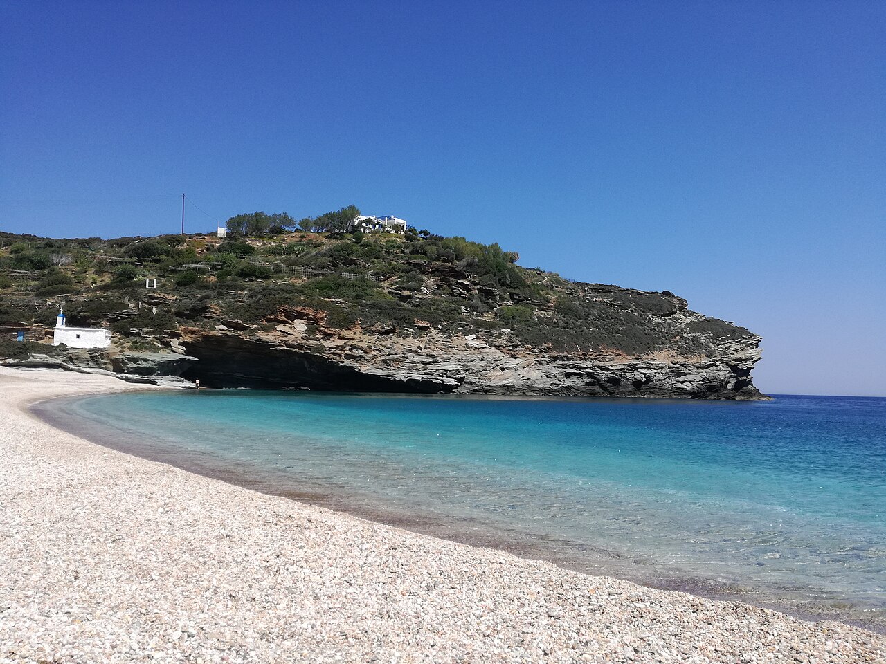 A crescent of pale pebbles and clear turquoise water below a rocky headland, with a small white chapel above the shore.