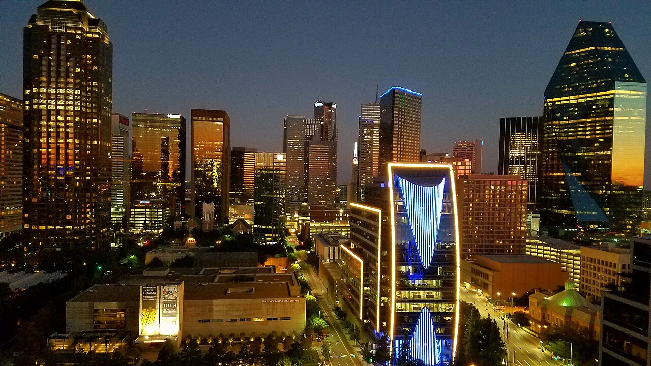 A nighttime view of the Dallas skyline with illuminated skyscrapers and a bright blue building.