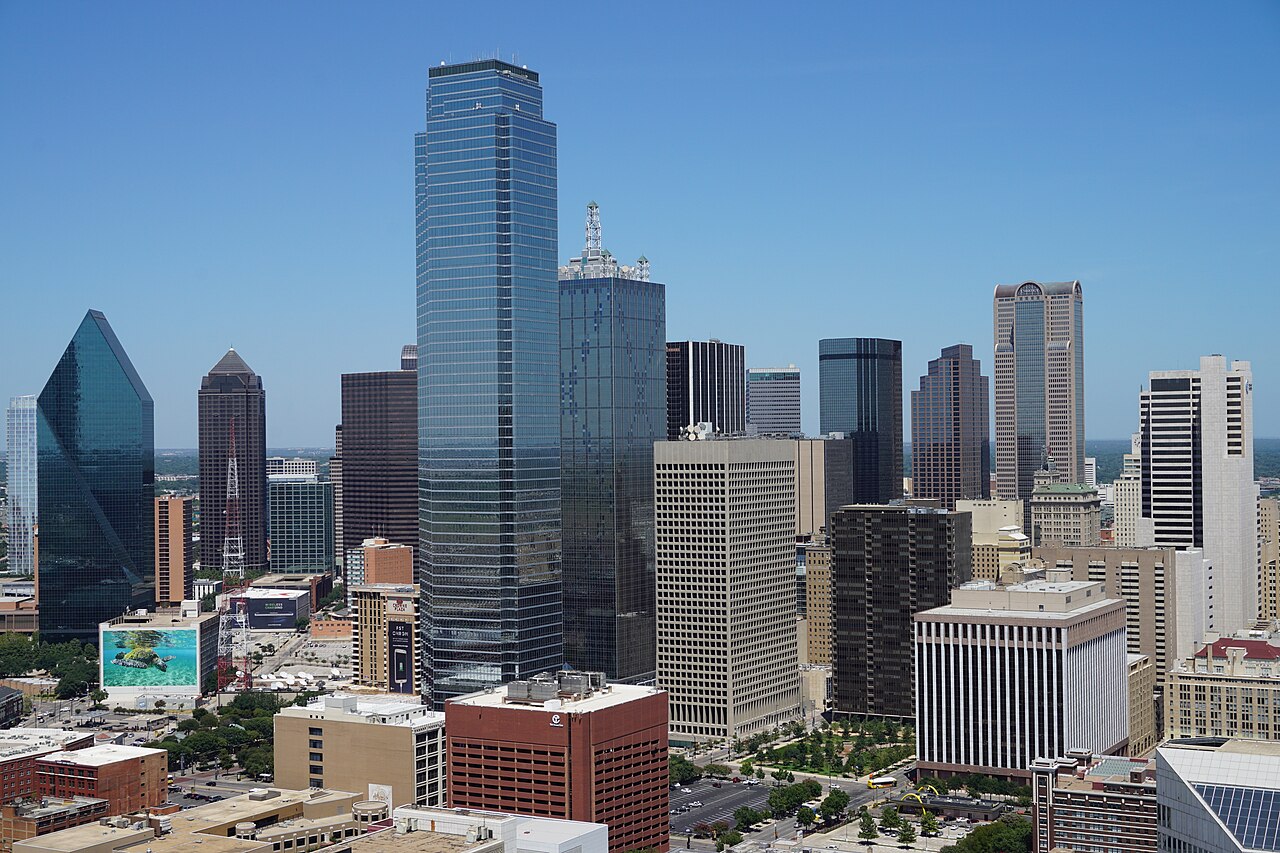 A wide shot of the Dallas skyline with numerous skyscrapers under a clear blue sky.