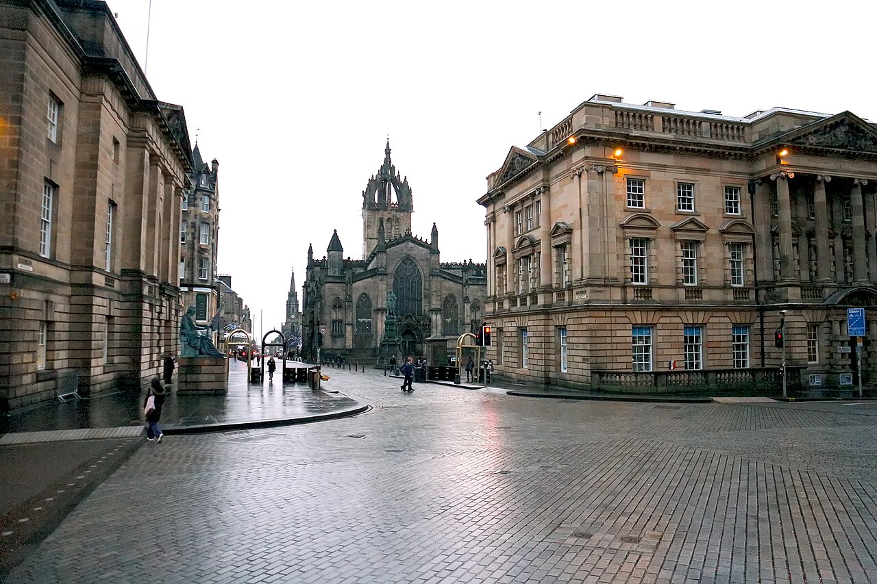 A wet paved street between grand stone buildings, leading to a dark Gothic church with a crown-shaped spire.
