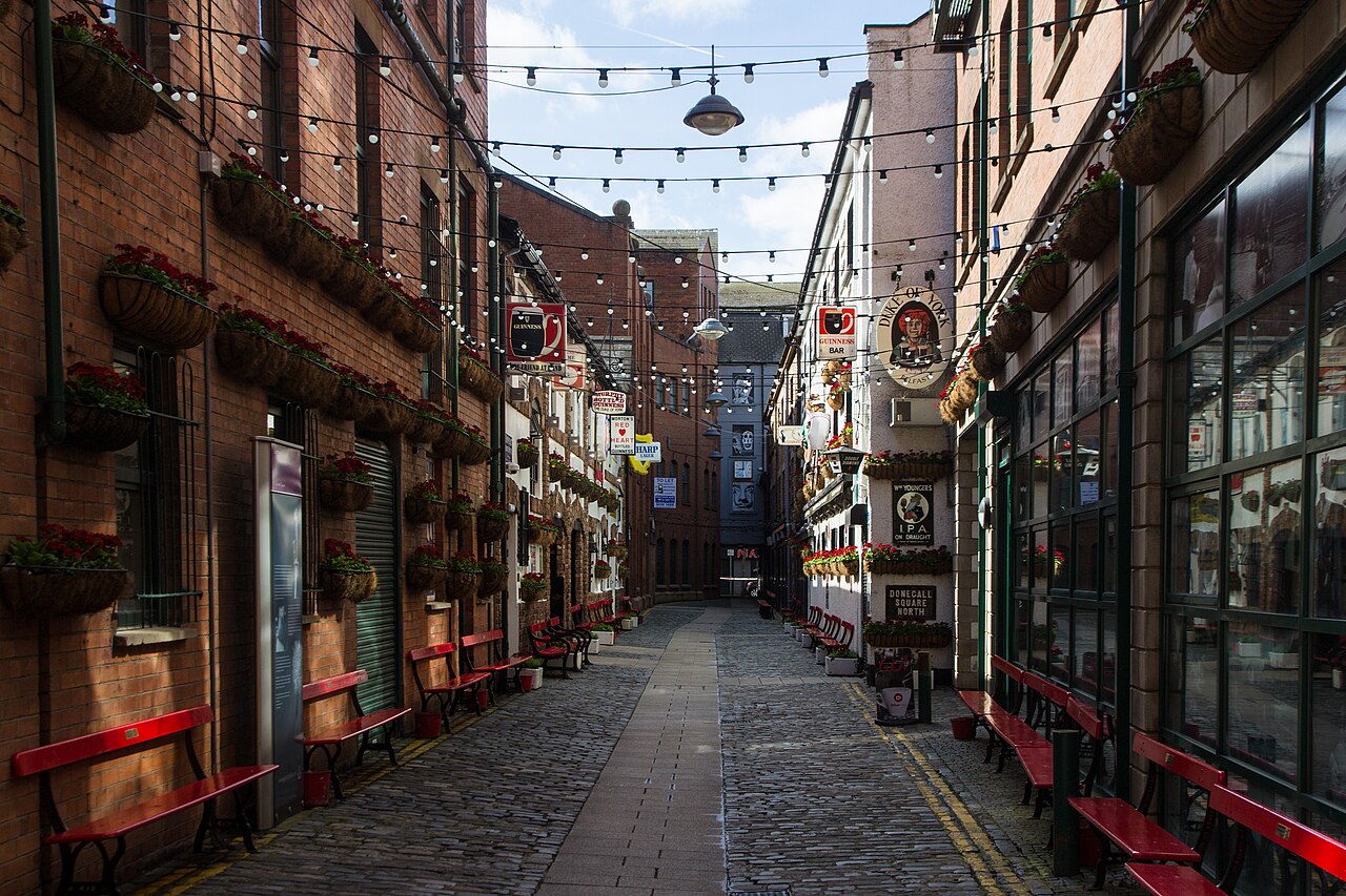 A narrow stone-paved alley between red-brick buildings, lined with red benches and hanging baskets, with strings of lights overhead.