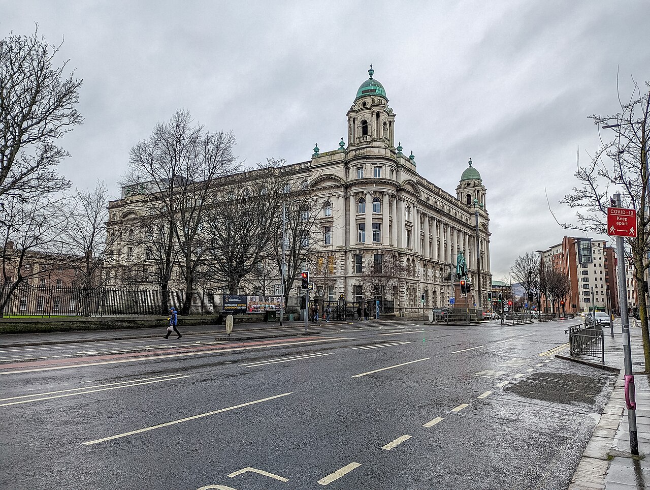 A grand stone civic building with a green copper dome and corner towers behind bare winter trees, across a wet, empty road.