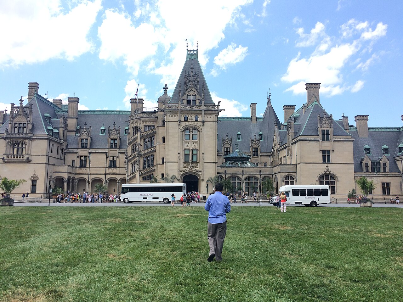 The grand facade of a large chateau-style building with many windows and turrets, with people and tour buses in front.