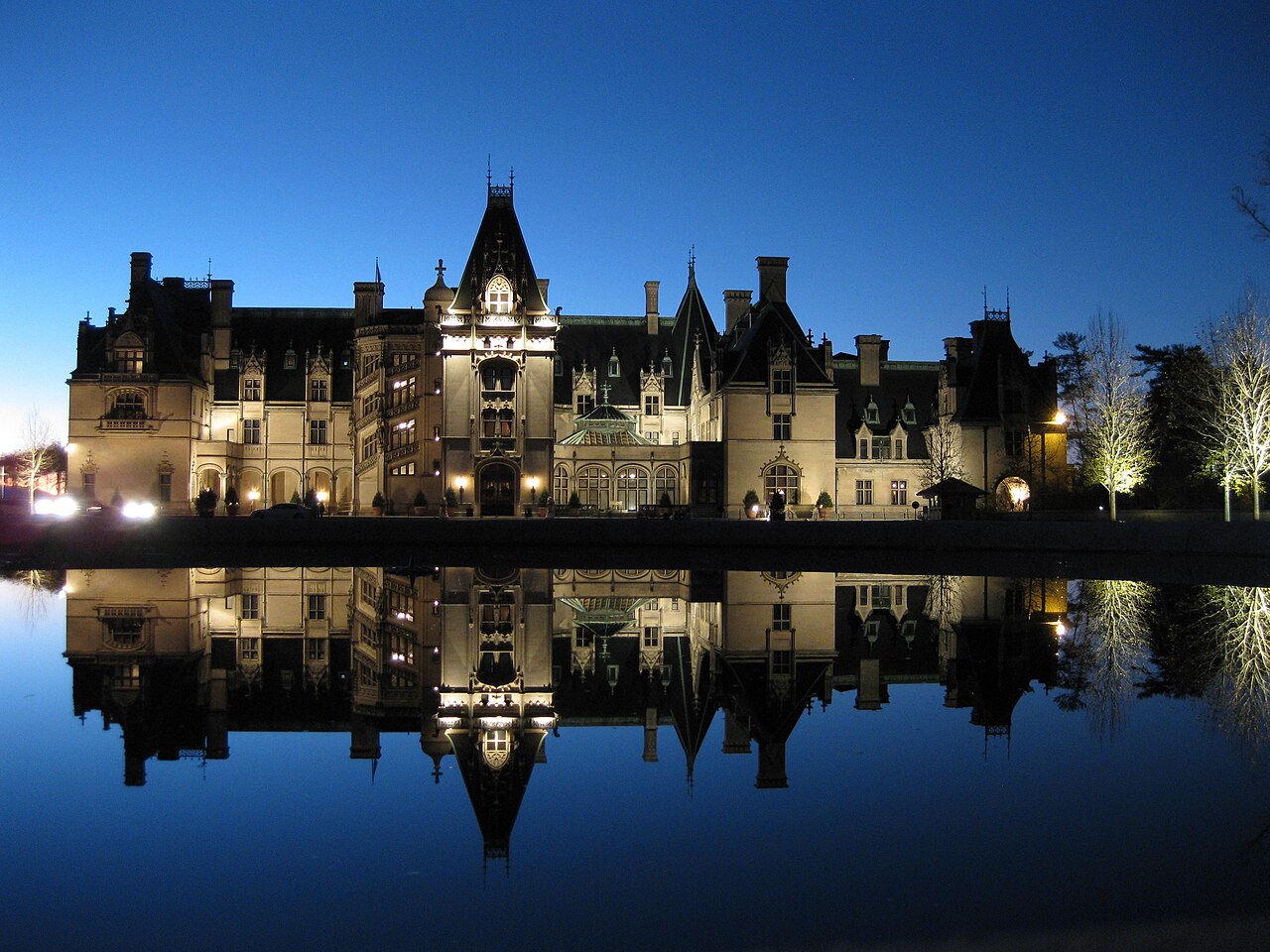 A large, ornate mansion with multiple turrets and chimneys is reflected in a still body of water under a dark blue sky.