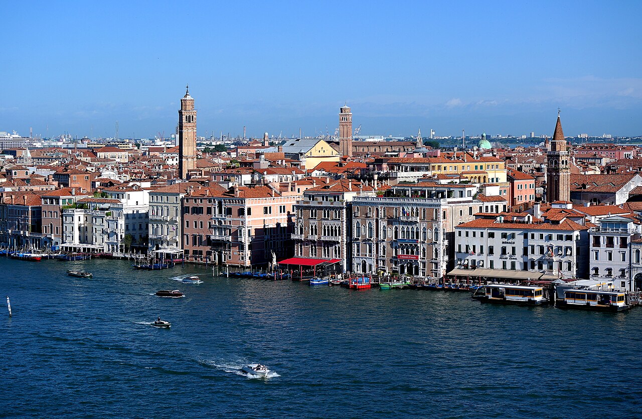 Seen from above, palaces and hotels line a waterfront busy with boats, with red rooftops and bell towers stretching to the horizon.