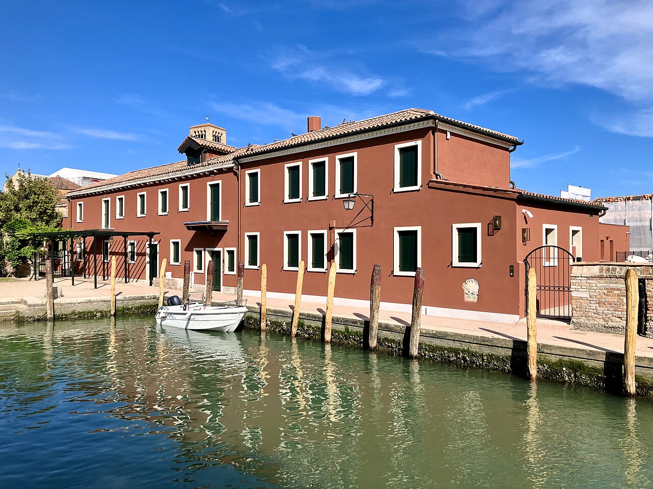 A long two-storey brick-red building with a tiled roof on a canal bank, with wooden mooring posts and a small boat on the green water.