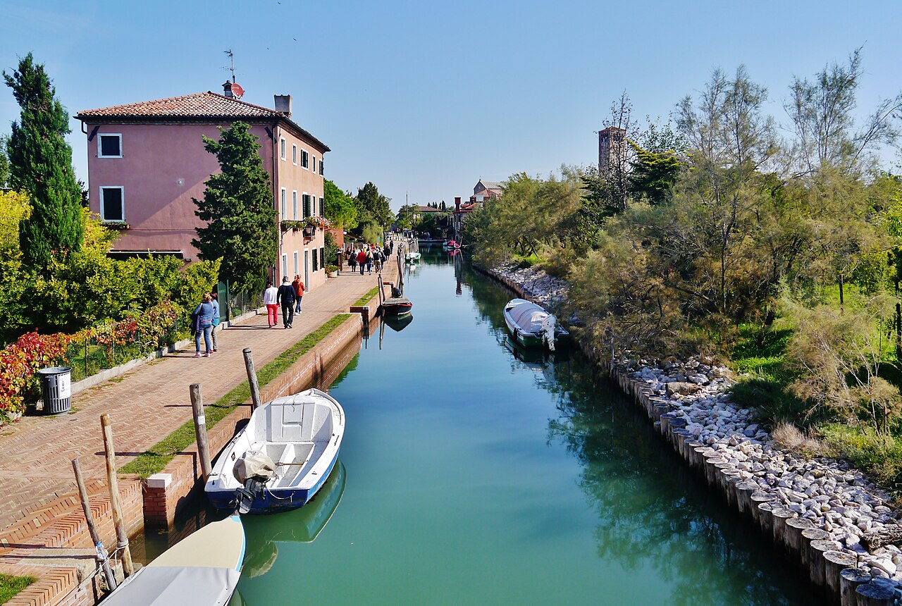 A narrow green canal with a brick path on the left, a pink house, moored boats and overgrown banks on the right.