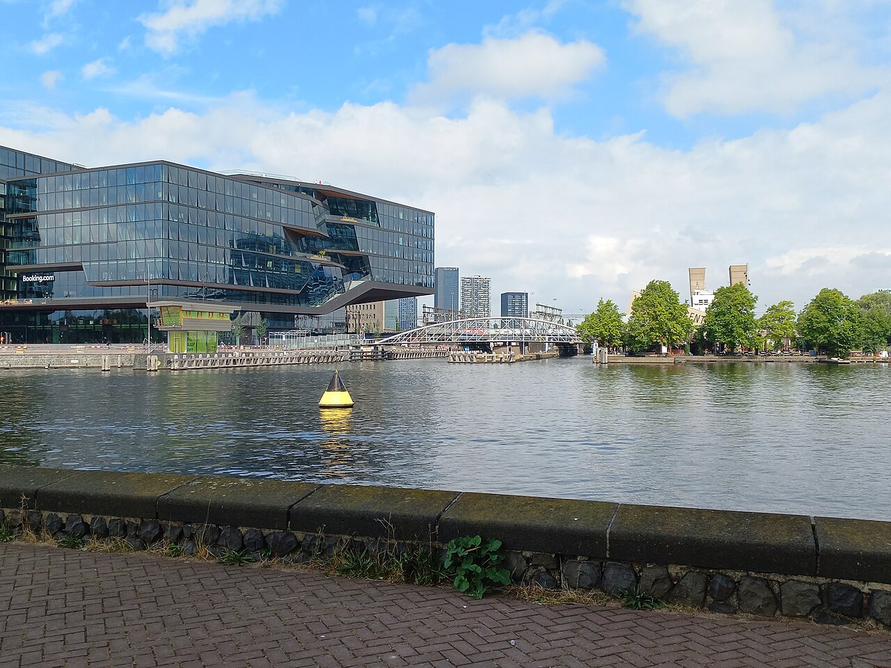 A dark glass office block stands across a stretch of calm water, with trees and a low bridge to its right.