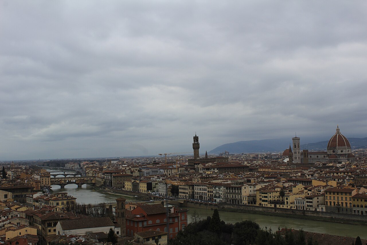 A wide shot of the Florence cityscape under a cloudy sky, with the Arno River winding through and historical buildings lining its banks.