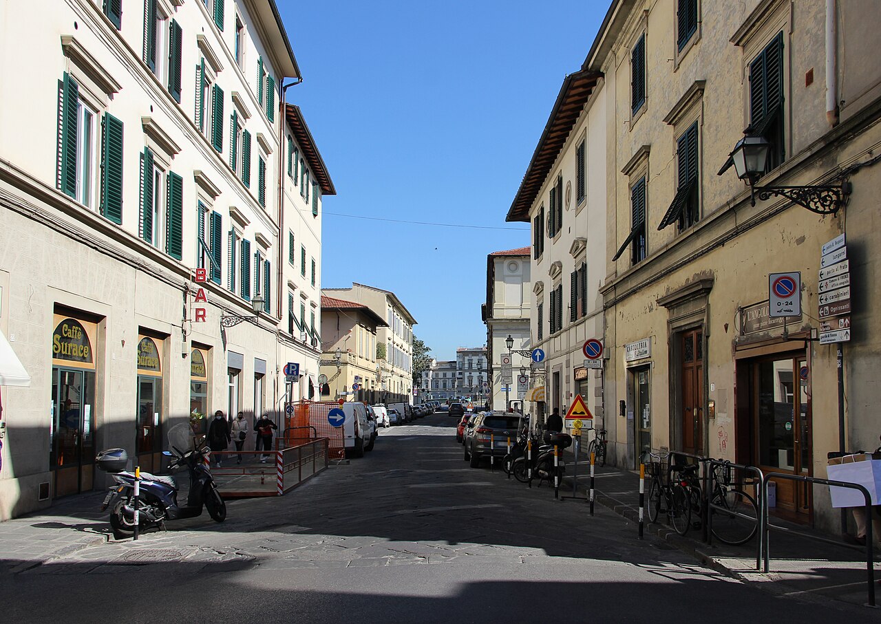 A street scene in Florence with buildings lining both sides of a paved road, parked cars, and a clear blue sky.