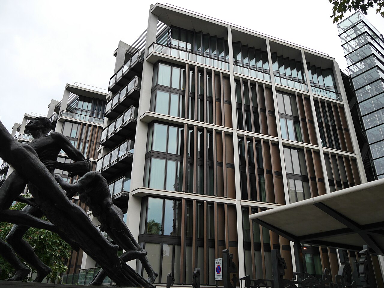 A modern apartment block of glass with slim vertical fins, seen from below, with the dark outline of a bronze sculpture in the foreground.