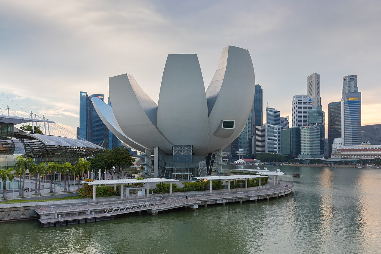 A white building shaped like an opening lotus flower on a waterfront, with a cluster of glass skyscrapers behind it in late-afternoon light.