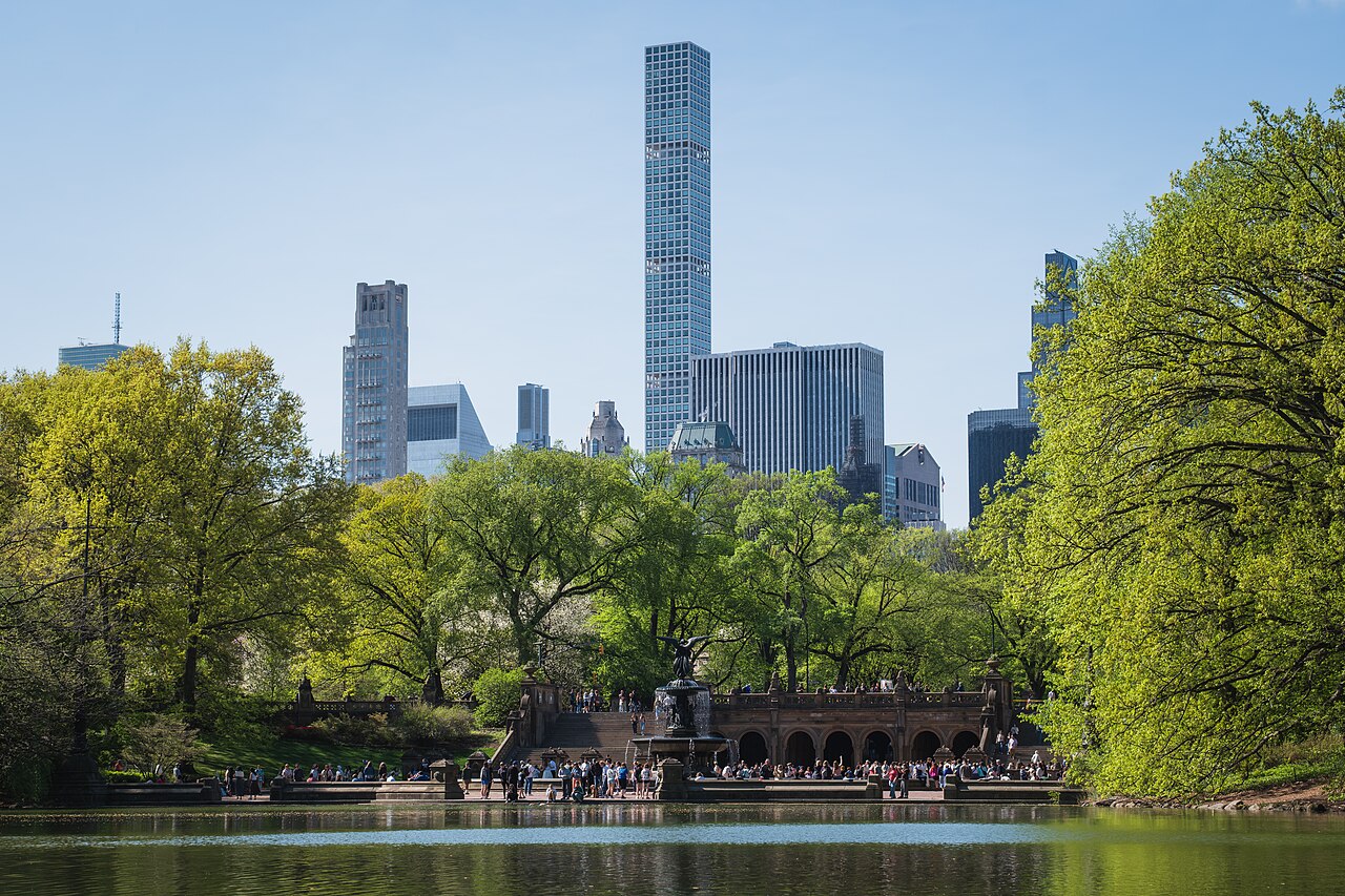 A lake ringed by spring trees with a stone terrace on the far shore, and a very tall, slender skyscraper rising above the skyline behind.