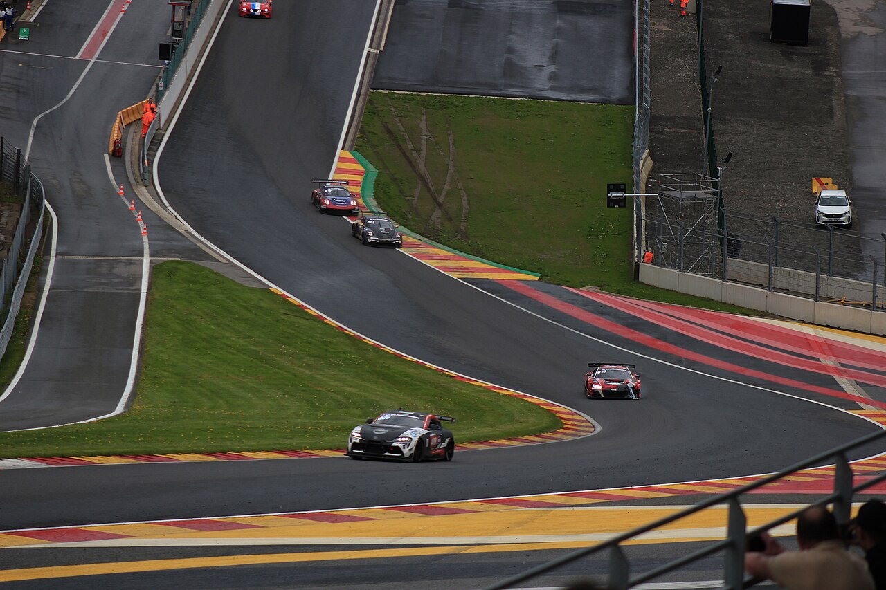 A steep uphill sweep of race track edged with red and yellow kerbs, with three racing cars climbing through the bend.