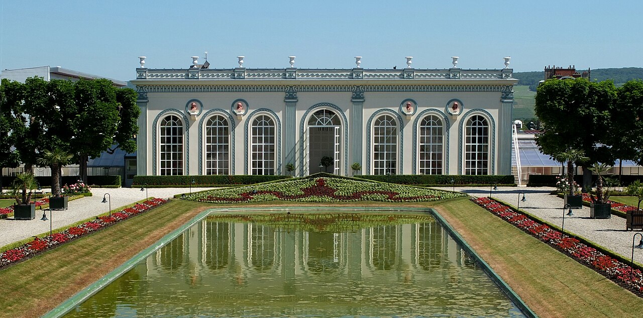 A long, low orangery with tall arched windows reflected in a rectangular pool, flanked by formal flower beds and clipped lawns.