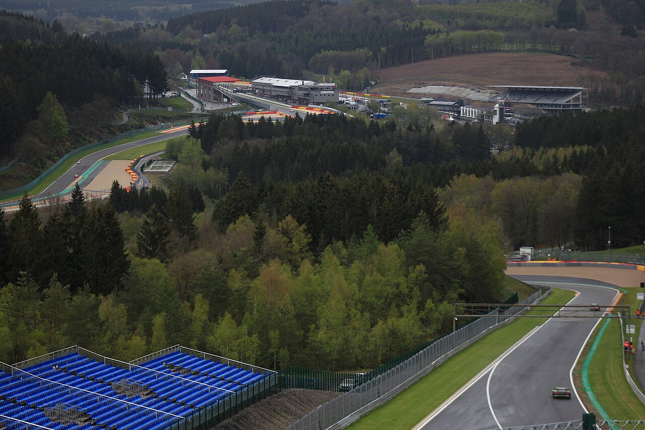 A race track winding through dense forest on a hillside, with rows of blue grandstand seats in the foreground and paddock buildings in the distance.