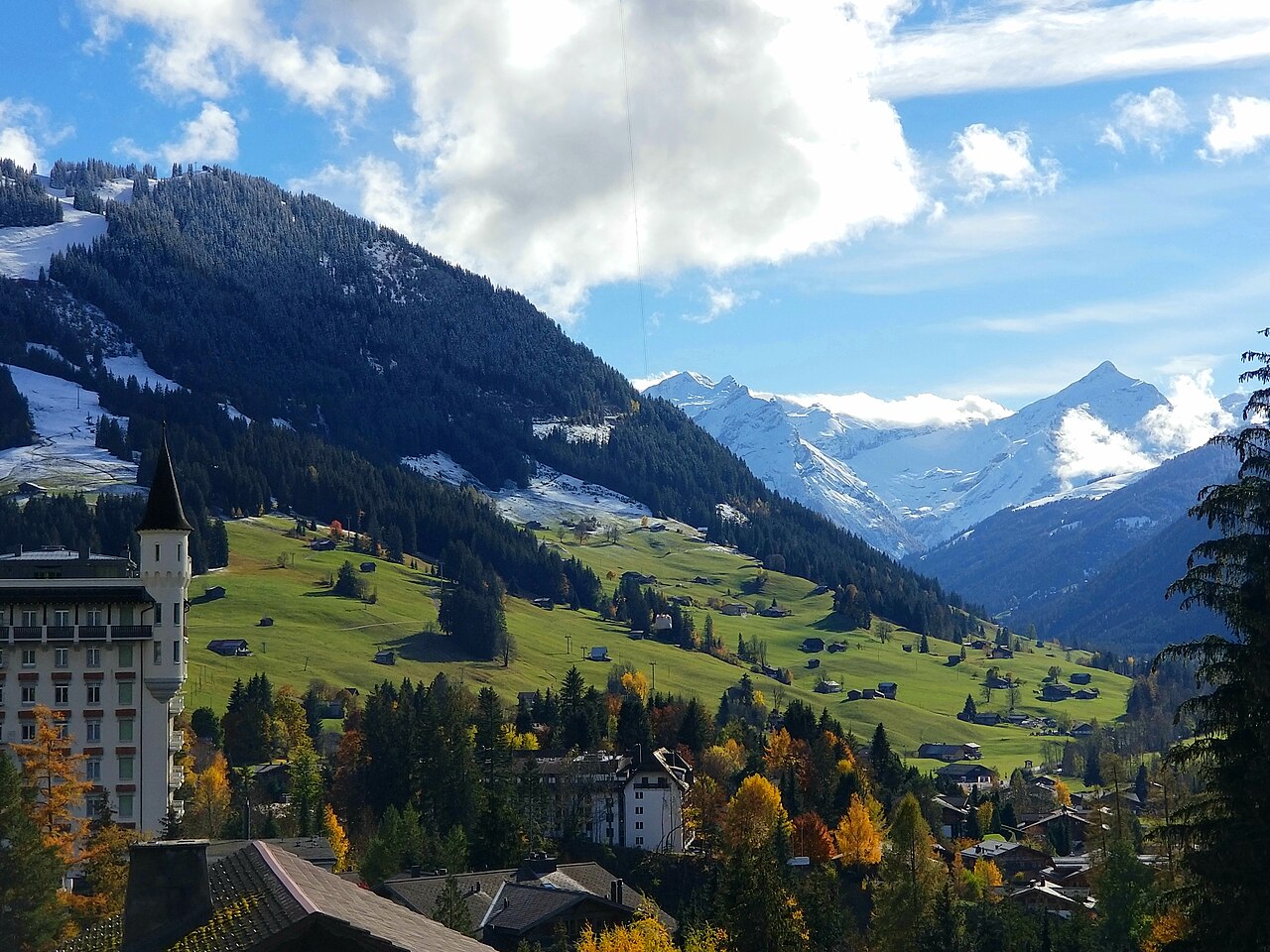A large building with a distinctive turret stands on a hillside, with a village below and snow-capped mountains in the distance.