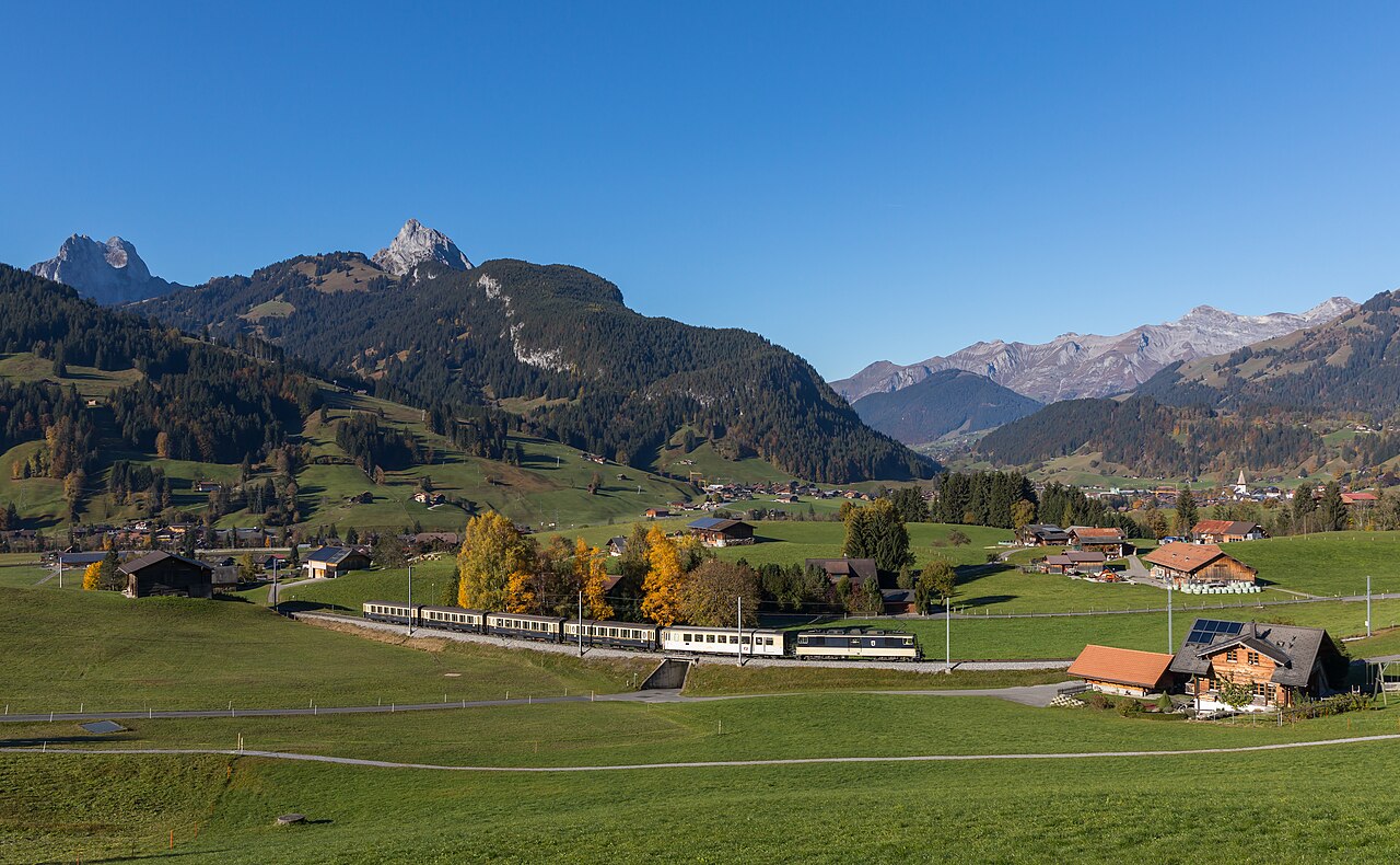 A train with several carriages moves through a green valley with mountains in the background under a clear blue sky.