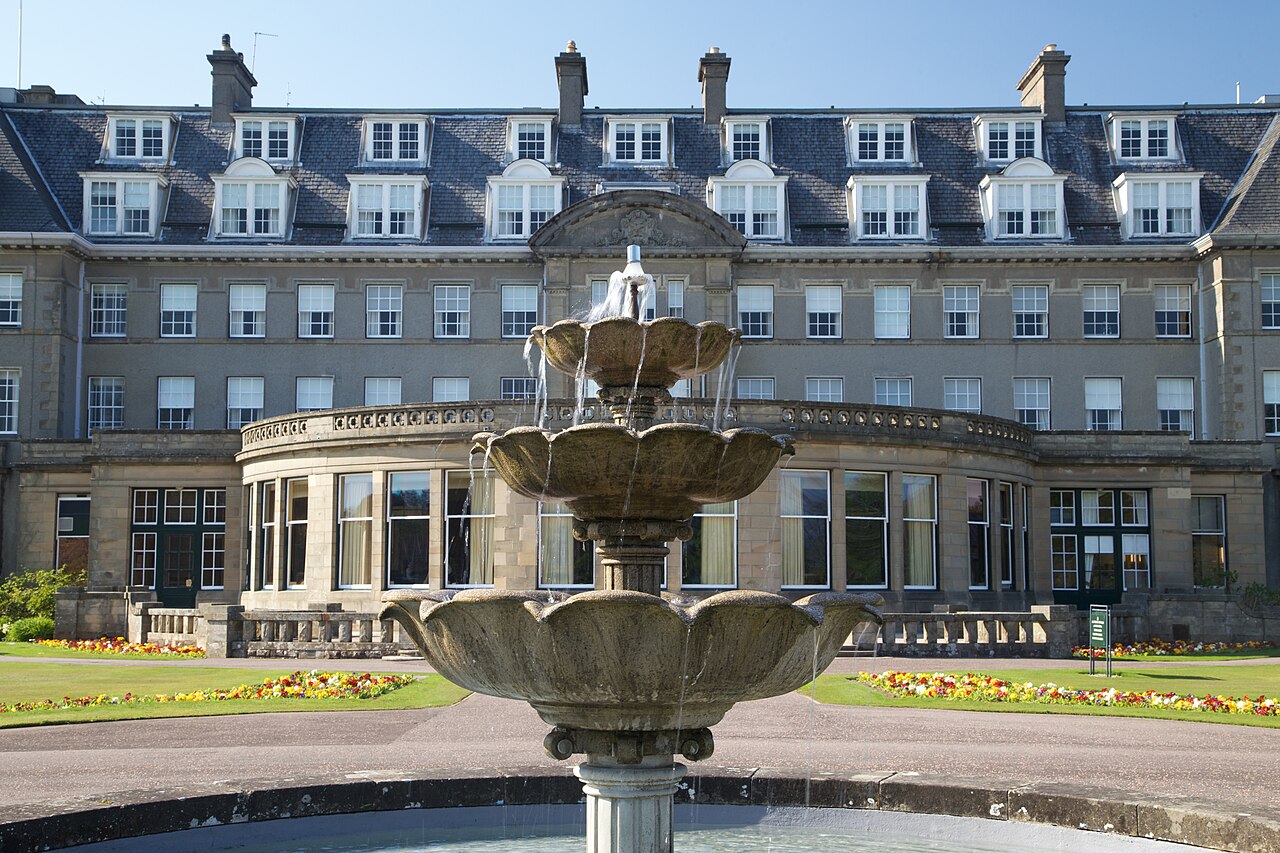 A long grey stone hotel with rows of sash windows and dormers stands behind a tiered stone fountain and a lawn.