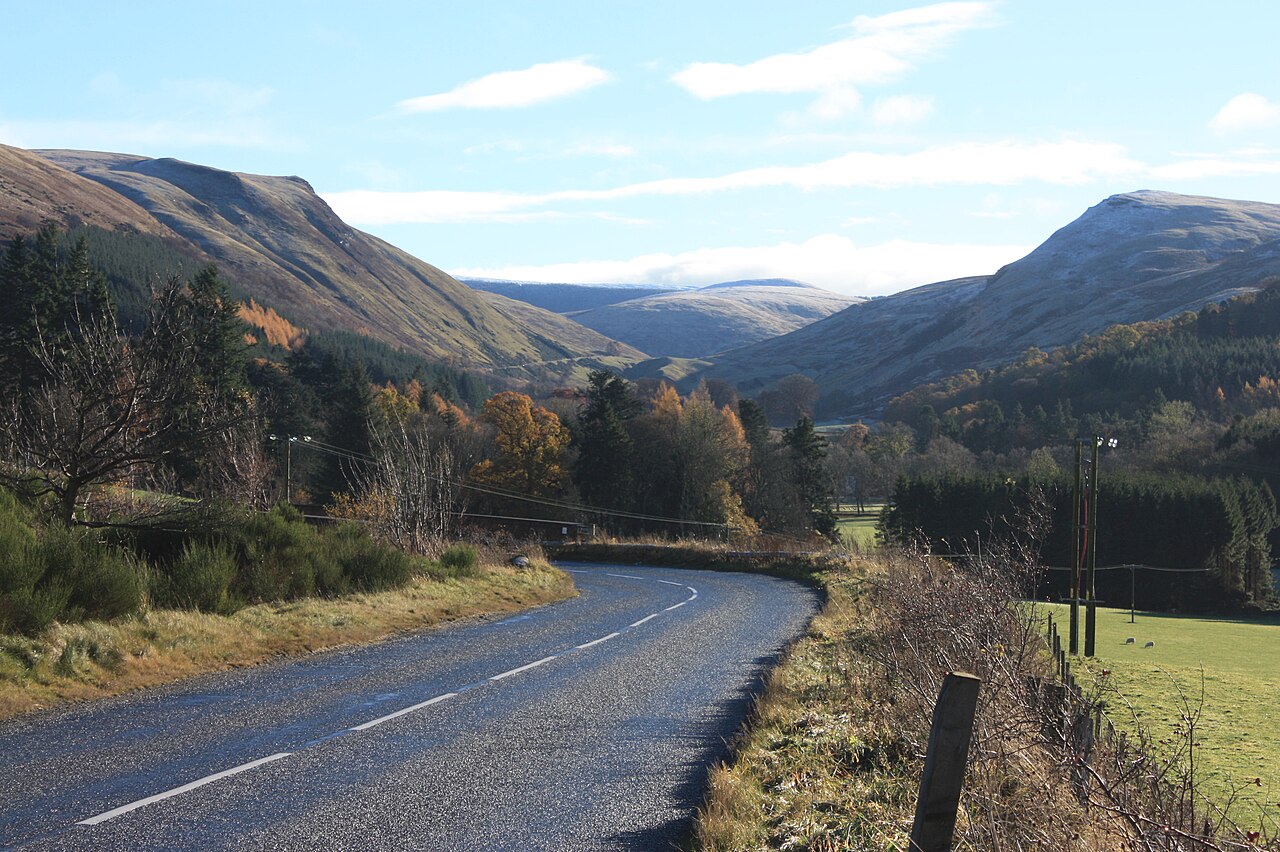 A road curves through a valley between steep brown and green hills under a blue sky.
