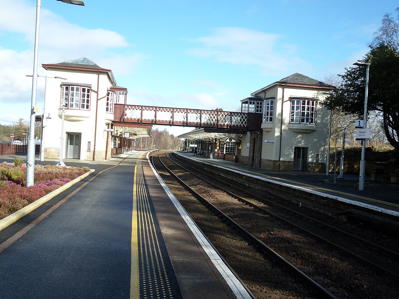 Two empty platforms flank a railway line, linked by a covered lattice footbridge between cream-painted station buildings.