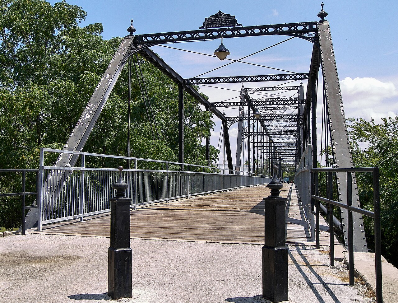 A steel truss bridge with a wooden deck and black railings stretches away between trees.