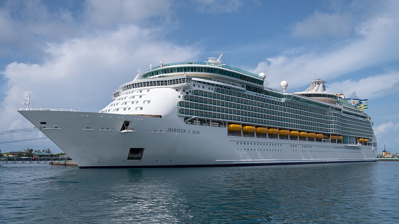 A large white cruise ship with many decks and yellow lifeboats docked in a harbor under a blue sky.