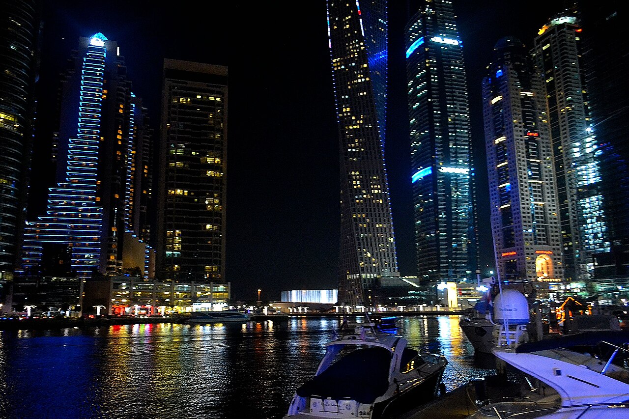 Illuminated skyscrapers are reflected in dark harbour water, with moored boats in the foreground.