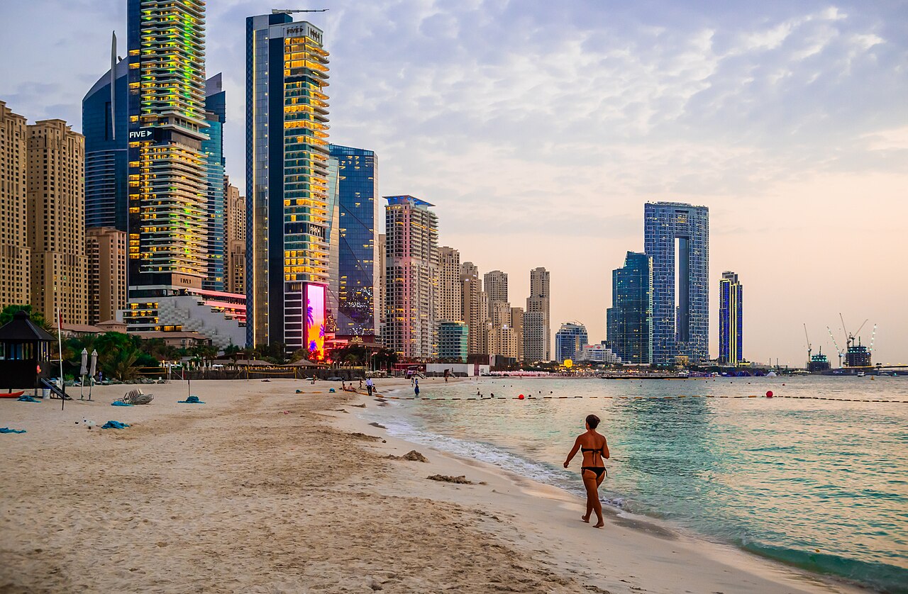 High-rise towers stand behind a sandy beach where a lone figure walks along the waterline.