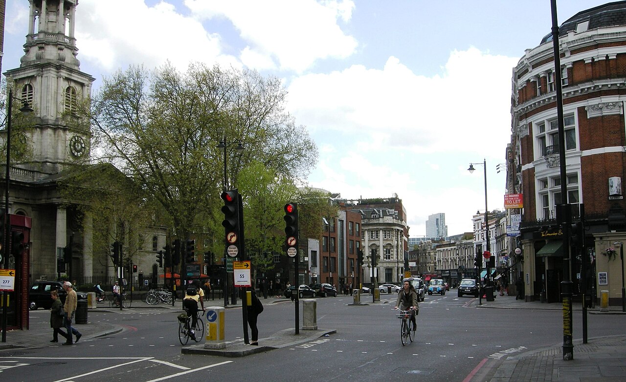 A wide city junction with traffic lights and a tree in leaf, a church portico on the left and brick buildings on the right.
