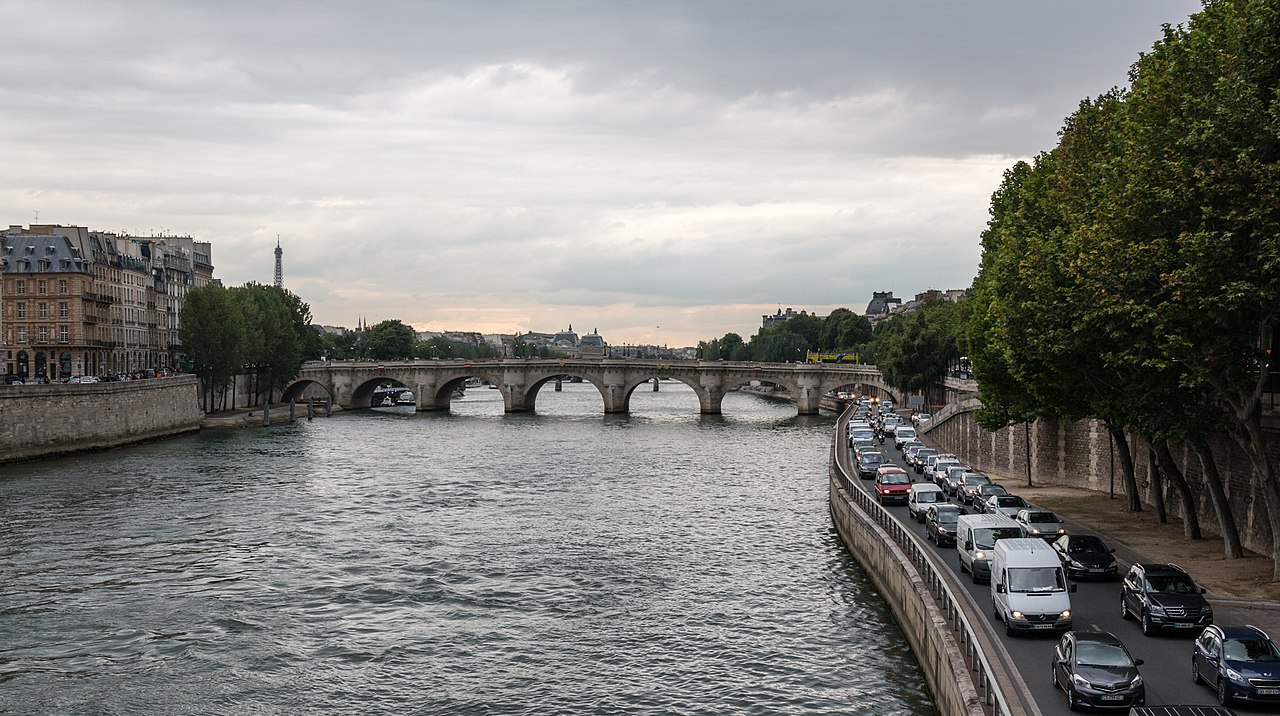 A wide river with a stone bridge and city buildings on both banks, with traffic on the right.