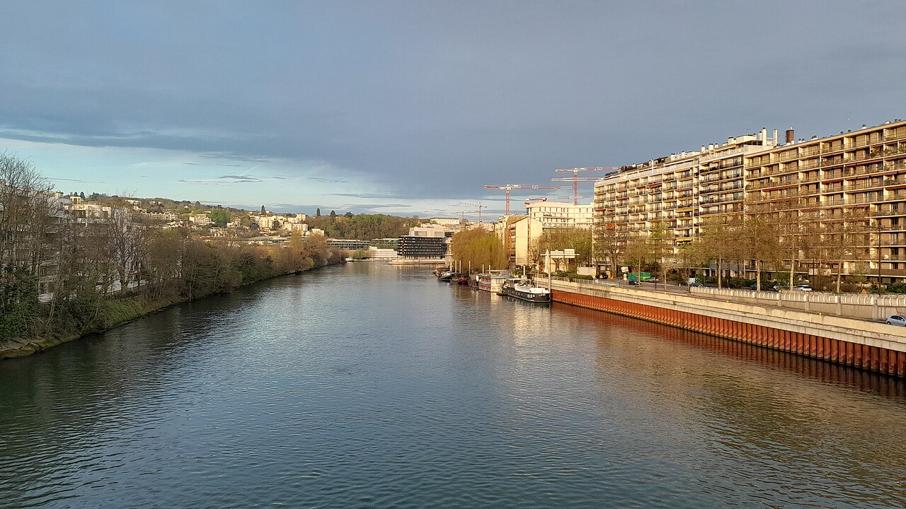 A wide river with buildings and trees on both banks under a cloudy sky at sunrise.