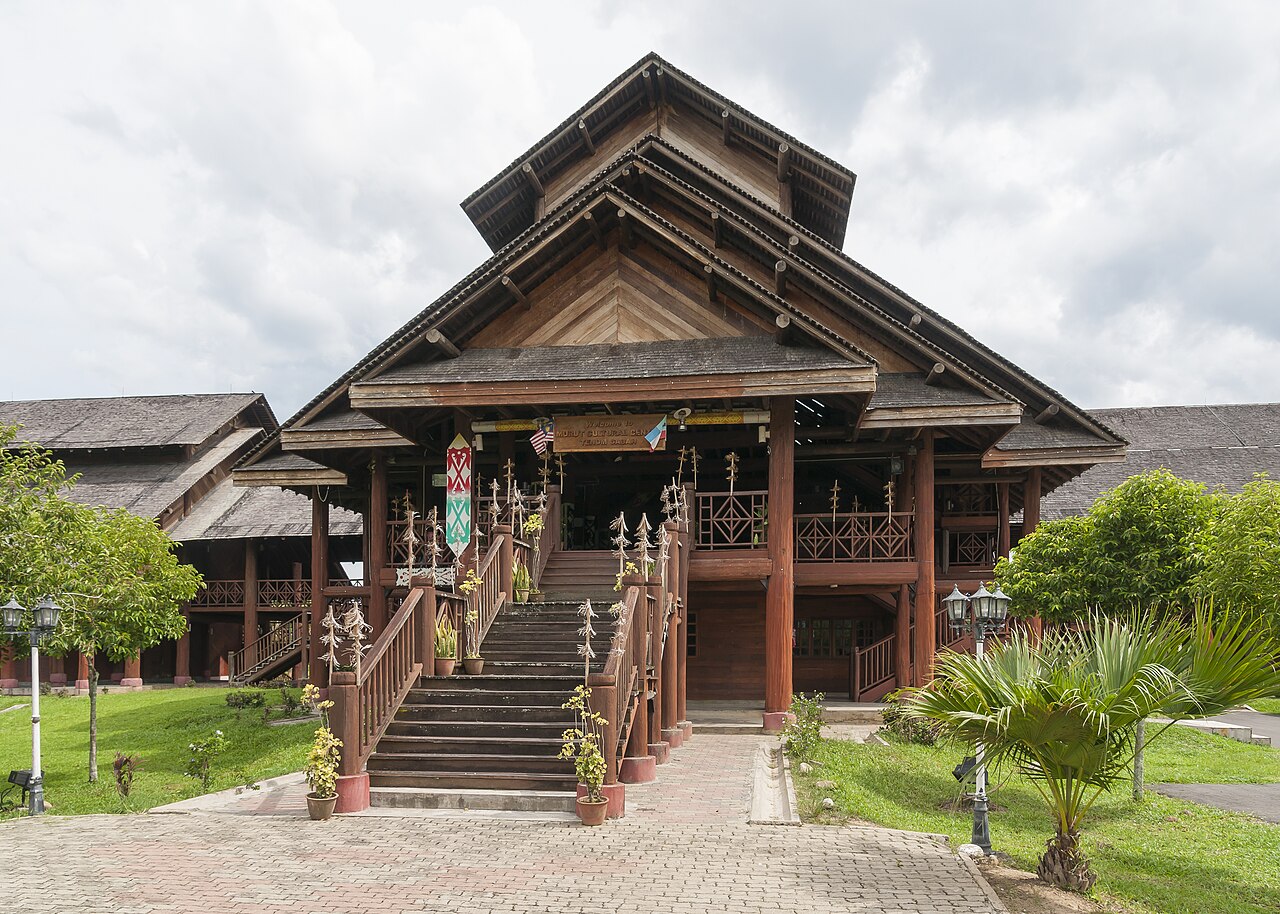A large, traditional wooden building with multiple tiered roofs and intricate carvings, surrounded by green grass and trees.
