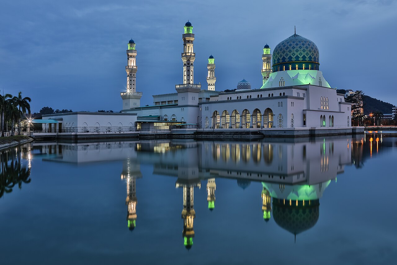 A large, ornate mosque with multiple domes and minarets, illuminated with green lights, reflected in still water under a twilight sky.