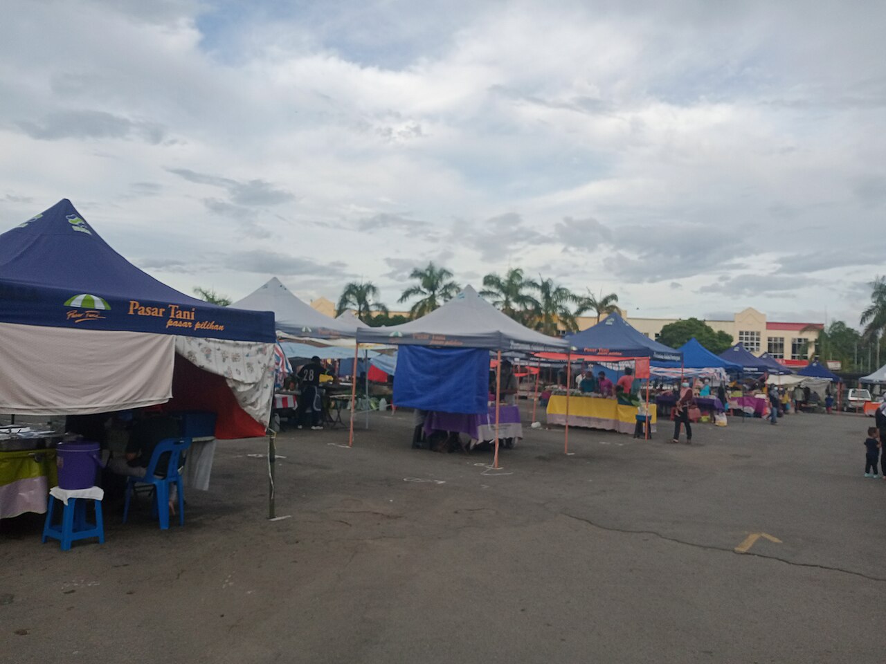 An outdoor market with numerous tents and stalls set up on a paved area under a cloudy sky, with buildings in the background.