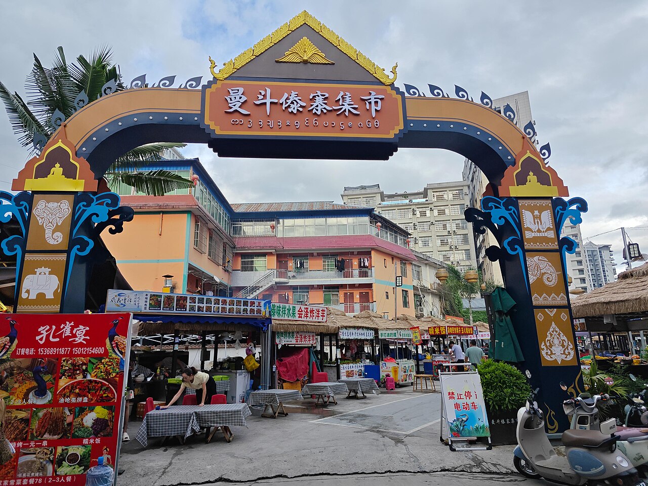 A large, ornate archway with traditional Dai architectural elements, leading into a bustling market area.