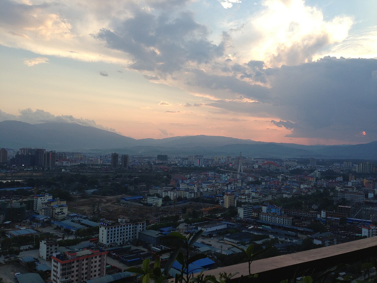An aerial view of a city with many buildings, surrounded by mountains under a cloudy sky at sunset.