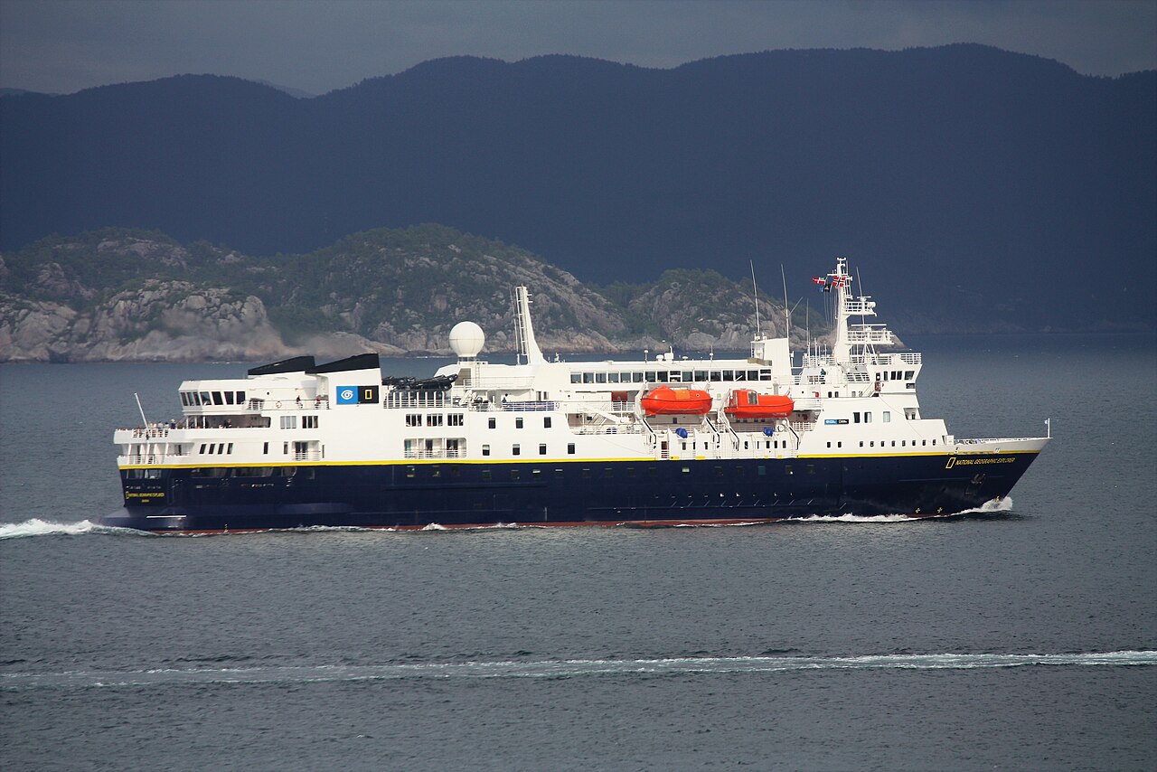 A white ship with a dark blue hull and orange lifeboats sails past dark, steep hills.