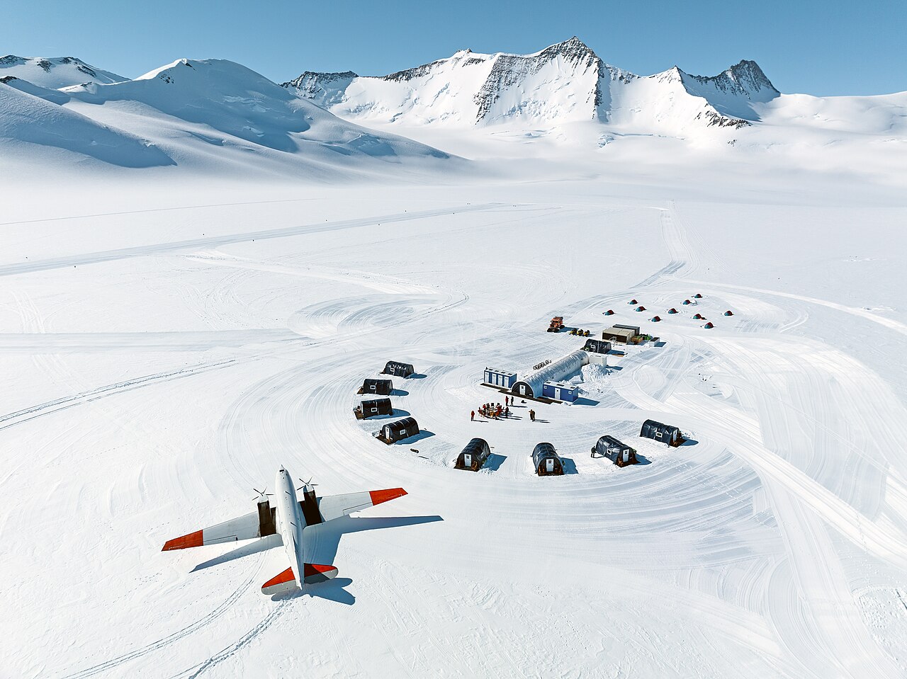 Seen from the air, a red-and-white propeller plane is parked on snow beside a cluster of small tents below white mountains.
