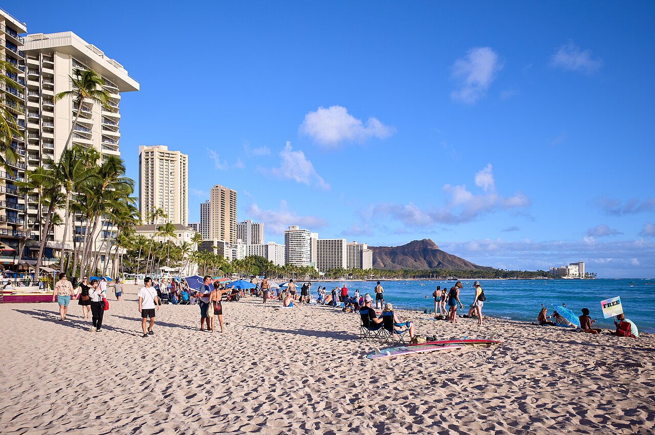 A wide sandy beach with sunbathers and palm trees, high-rise hotels along the shore and a volcanic ridge in the distance.