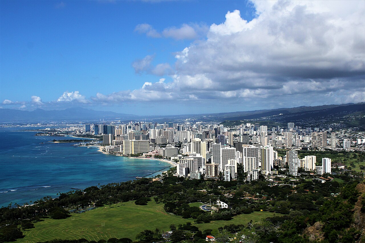A dense cluster of white high-rise towers beside a curving blue bay, with green mountains and tall clouds behind.