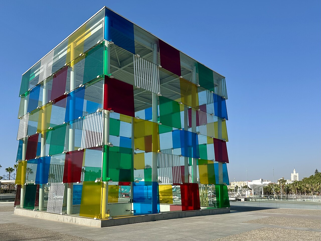 A large cube of clear and coloured glass panels in red, blue, yellow and green standing on an open paved plaza.