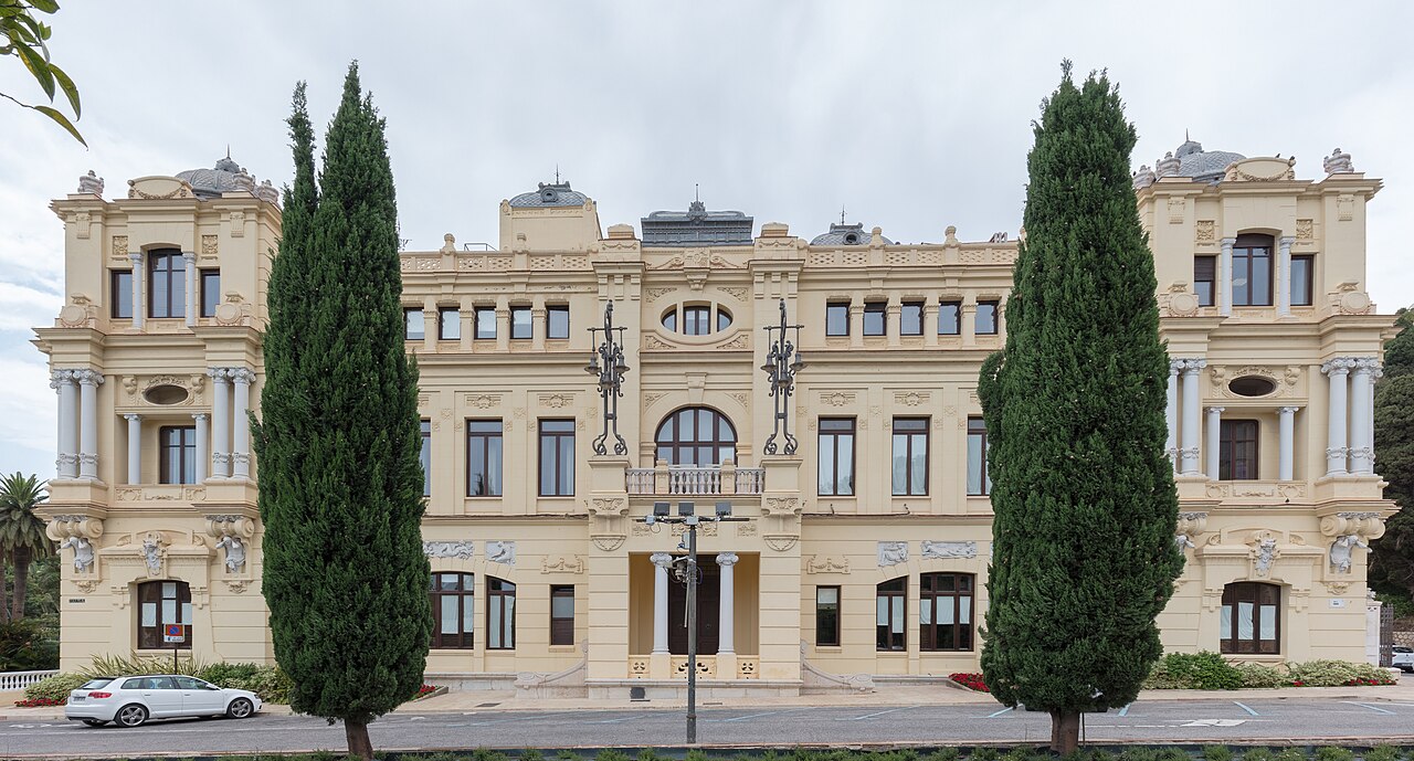 An ornate cream-coloured civic building with rows of tall windows and balconies, framed by two tall cypress trees.