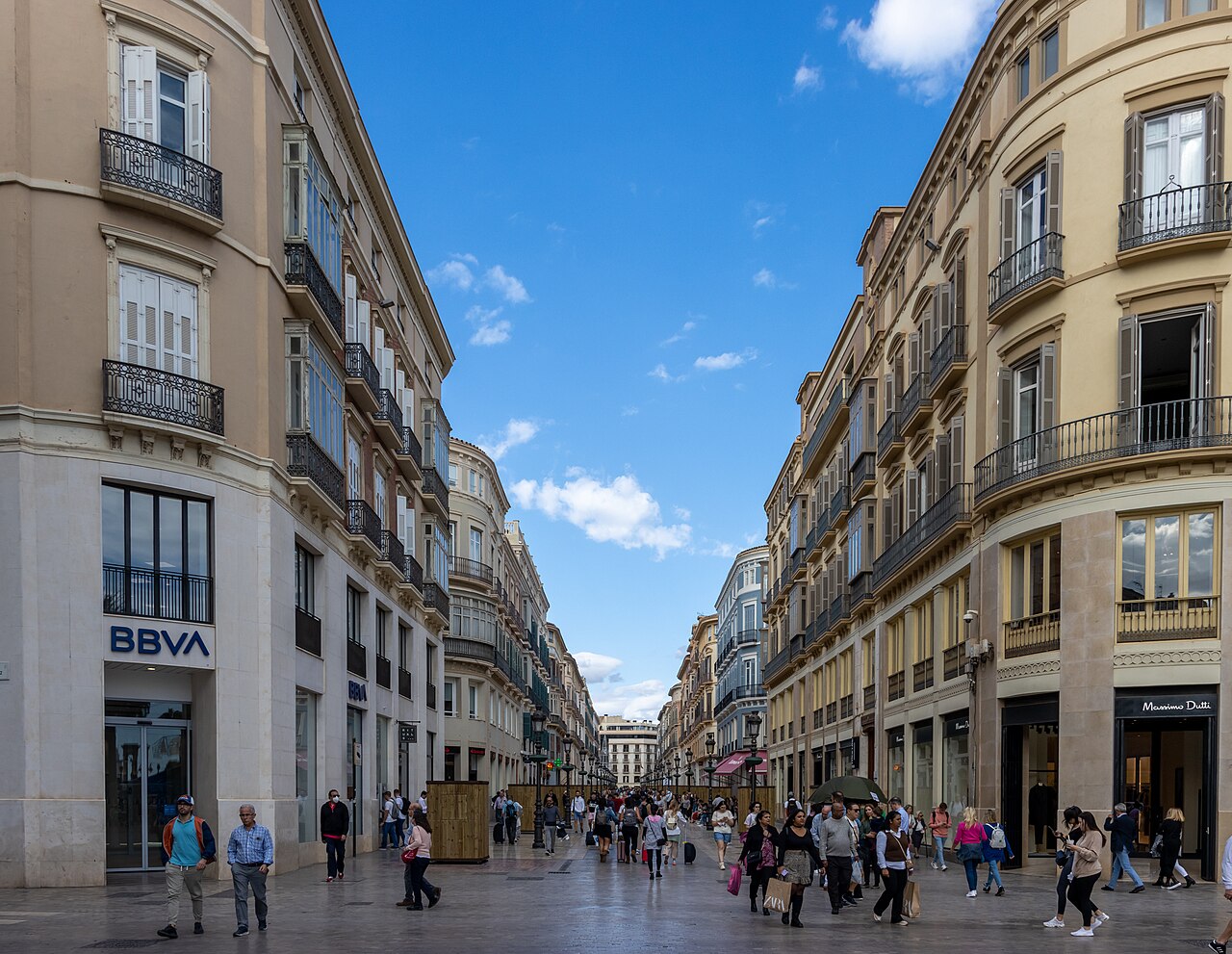 A straight pedestrian street lined with pale stone buildings with iron balconies, with shoppers walking in the distance.