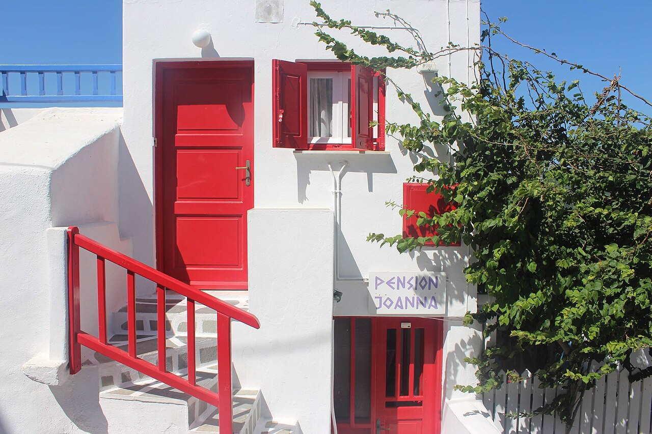 A white building with red doors and window shutters, a red railing, and green foliage.