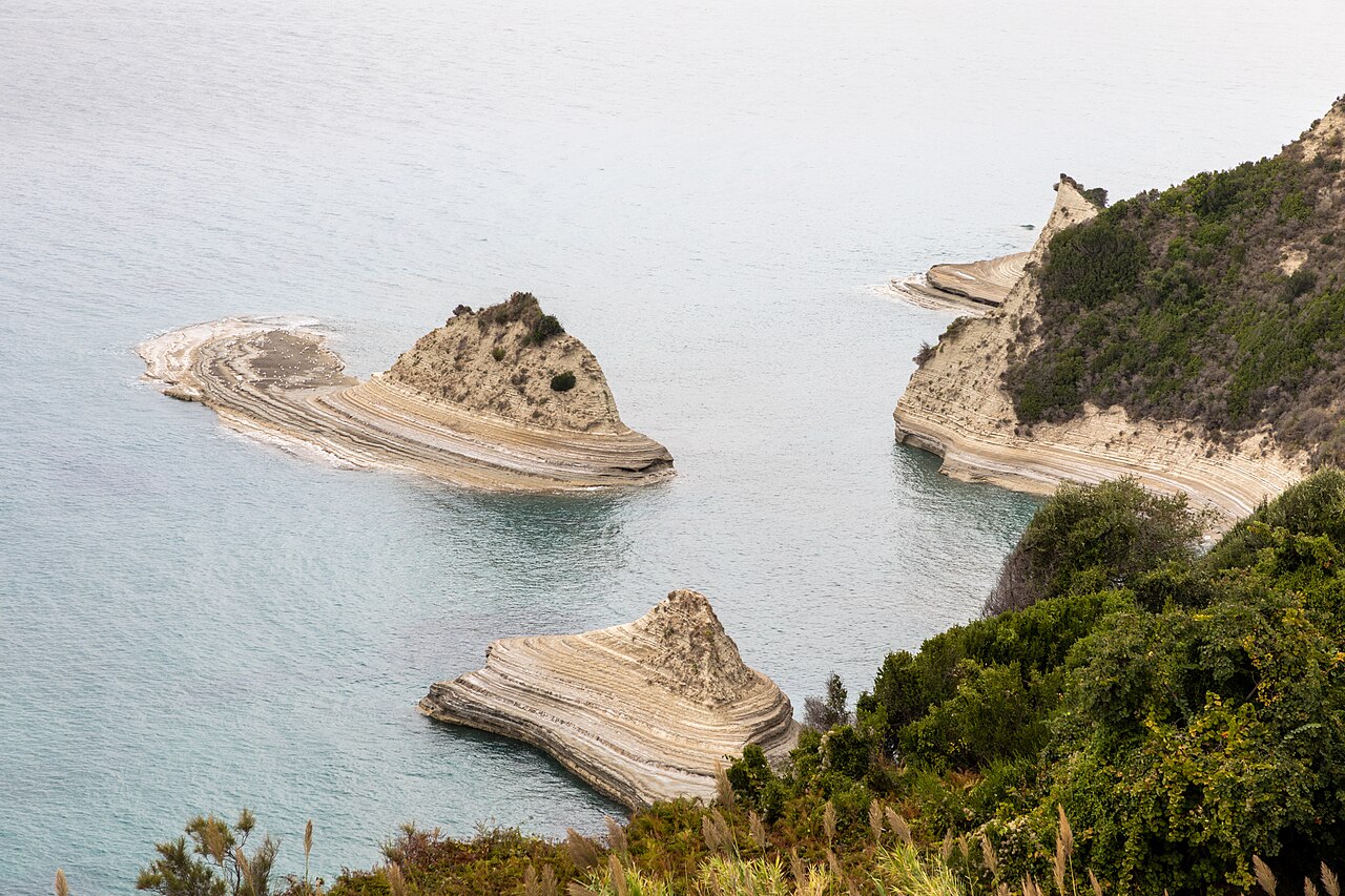 An aerial view of the coastline with layered rock formations extending into the clear blue-green sea.