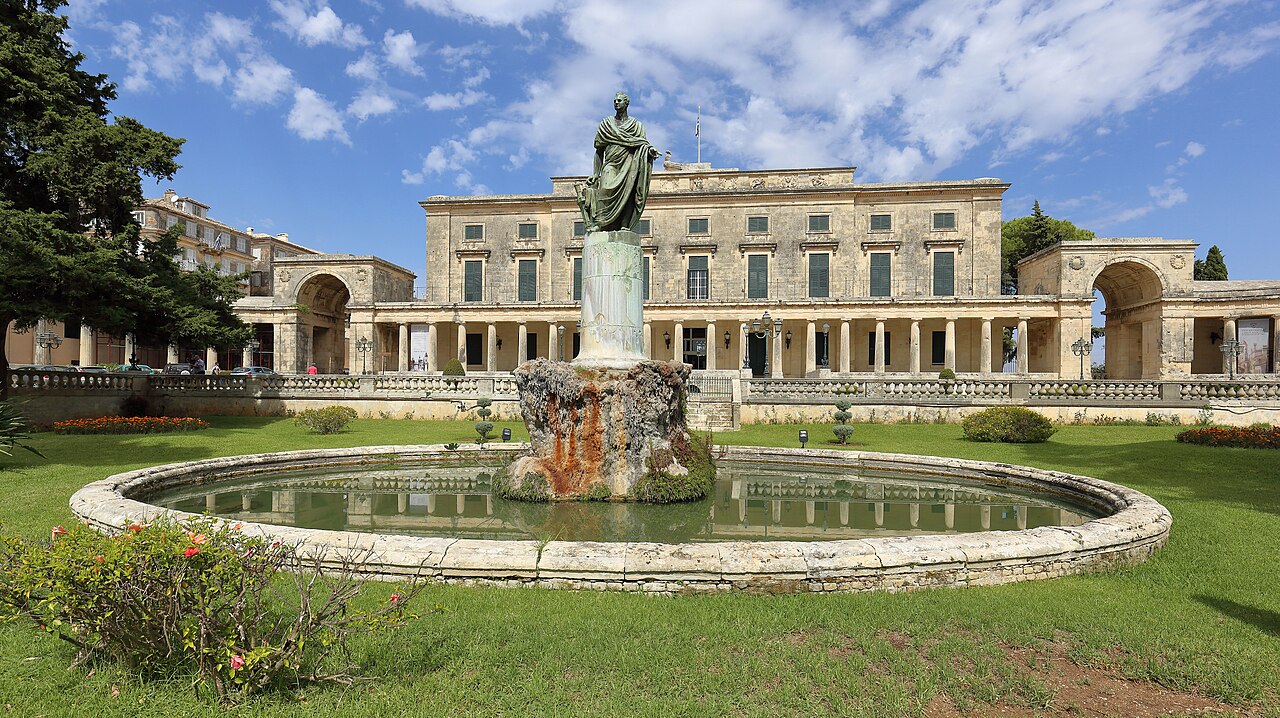 A grand neoclassical palace with a statue and fountain in front, surrounded by green lawns and trees under a blue sky.