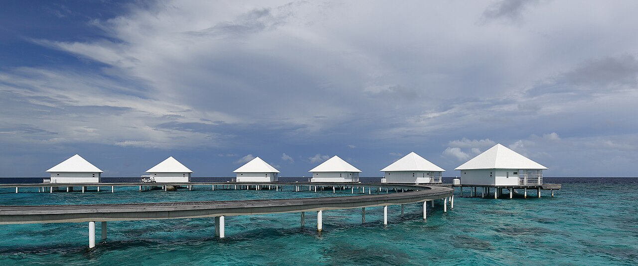 Overwater bungalows with white pyramid roofs extend along a curved wooden jetty over turquoise water under a cloudy sky.