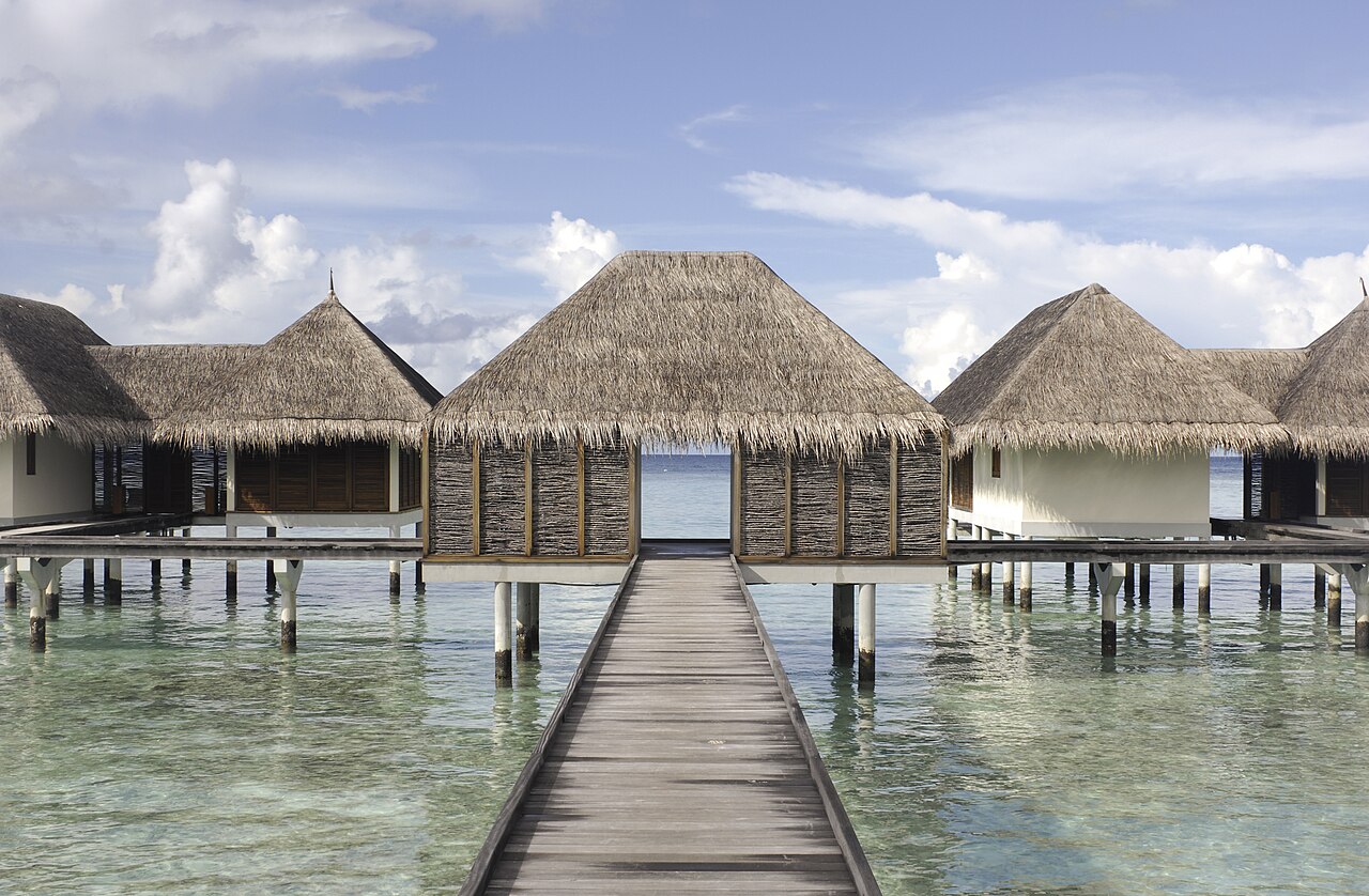 Overwater bungalows with thatched roofs are connected by a wooden pier over calm, clear water.
