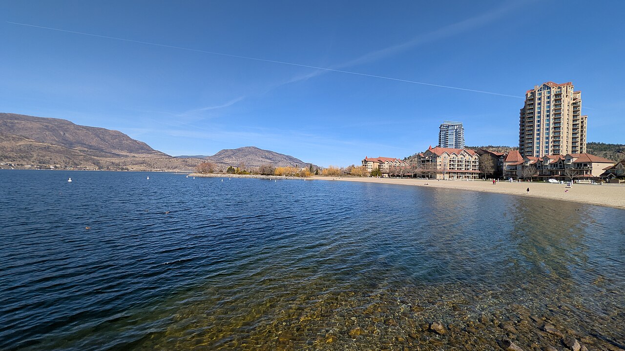 A wide shot of a clear lake with a sandy beach and several buildings, including a tall high-rise, along the shore, with mountains in the distance.
